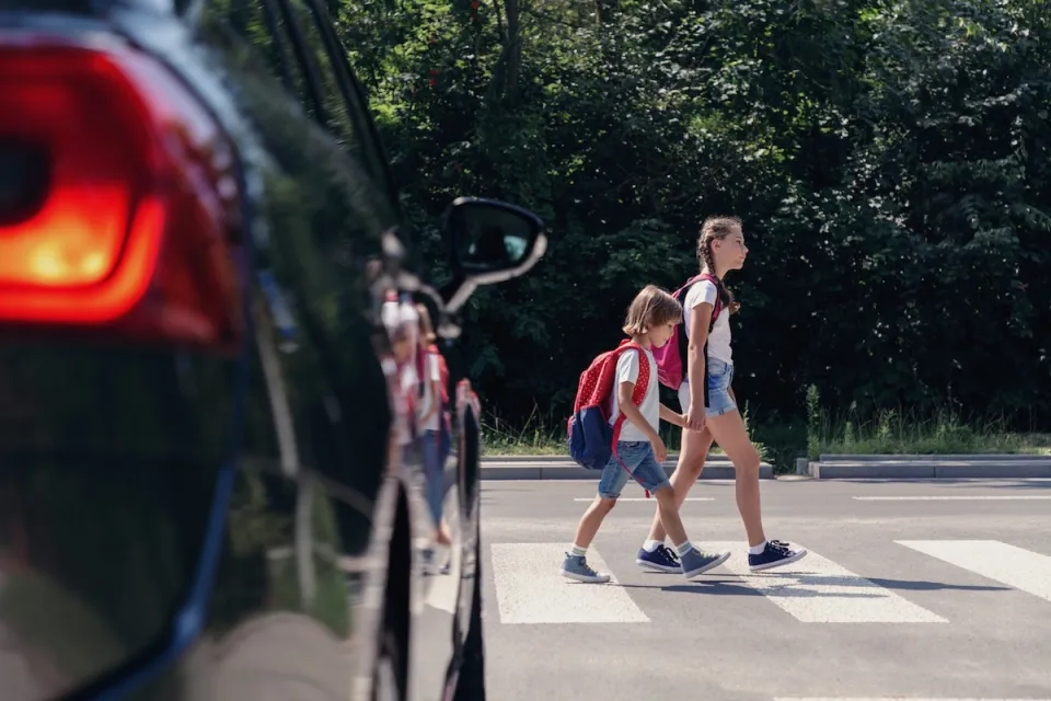 Two young children crossing street at a crosswalk in front of black car.